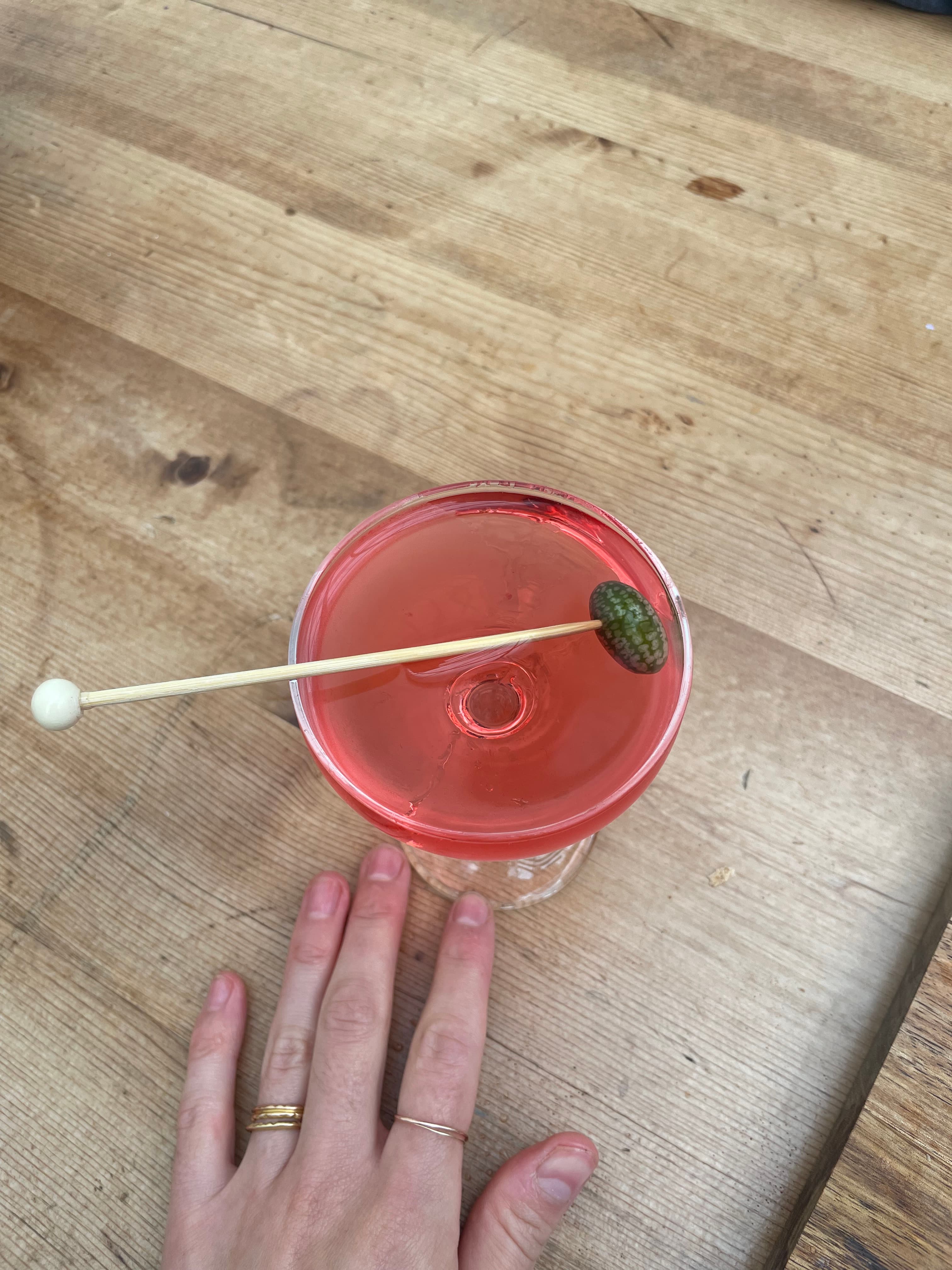 Pink cocktail garnished with a tiny cornichon on a wooden table