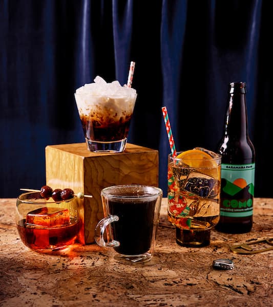 Group of four styled cocktails and a soda bottle on a cork surface