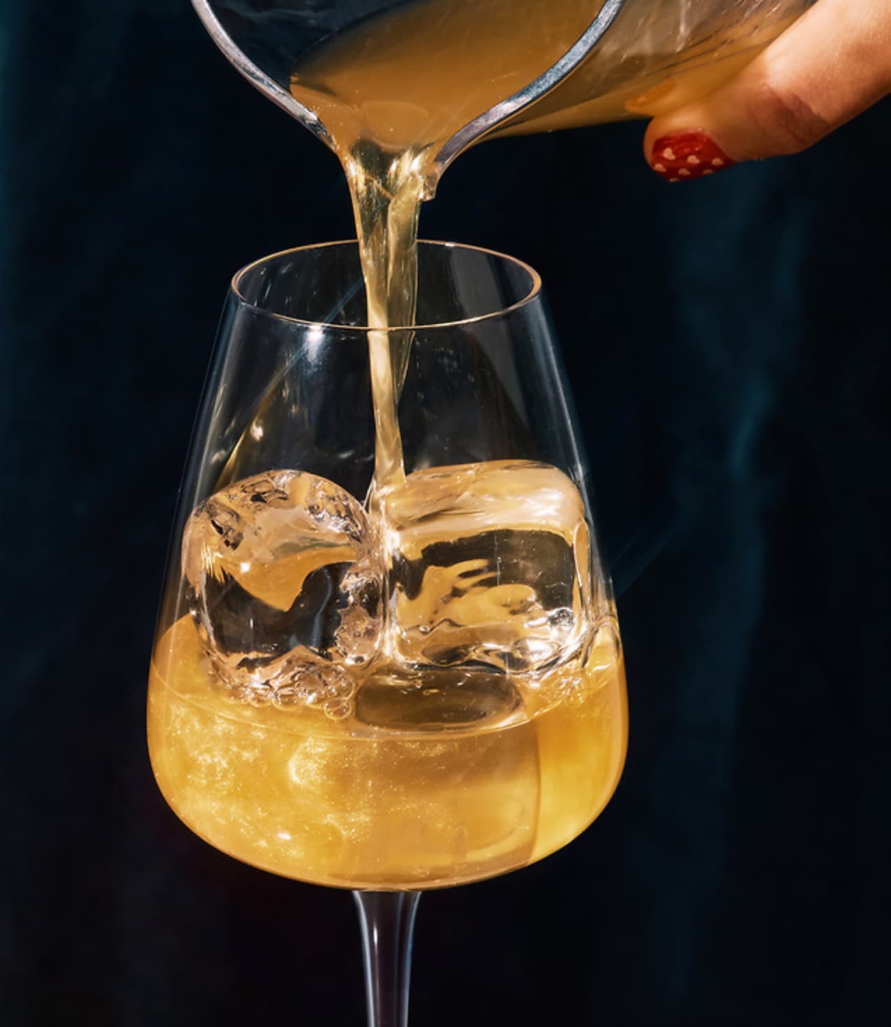 Golden cocktail being poured over clear ice in a wine glass