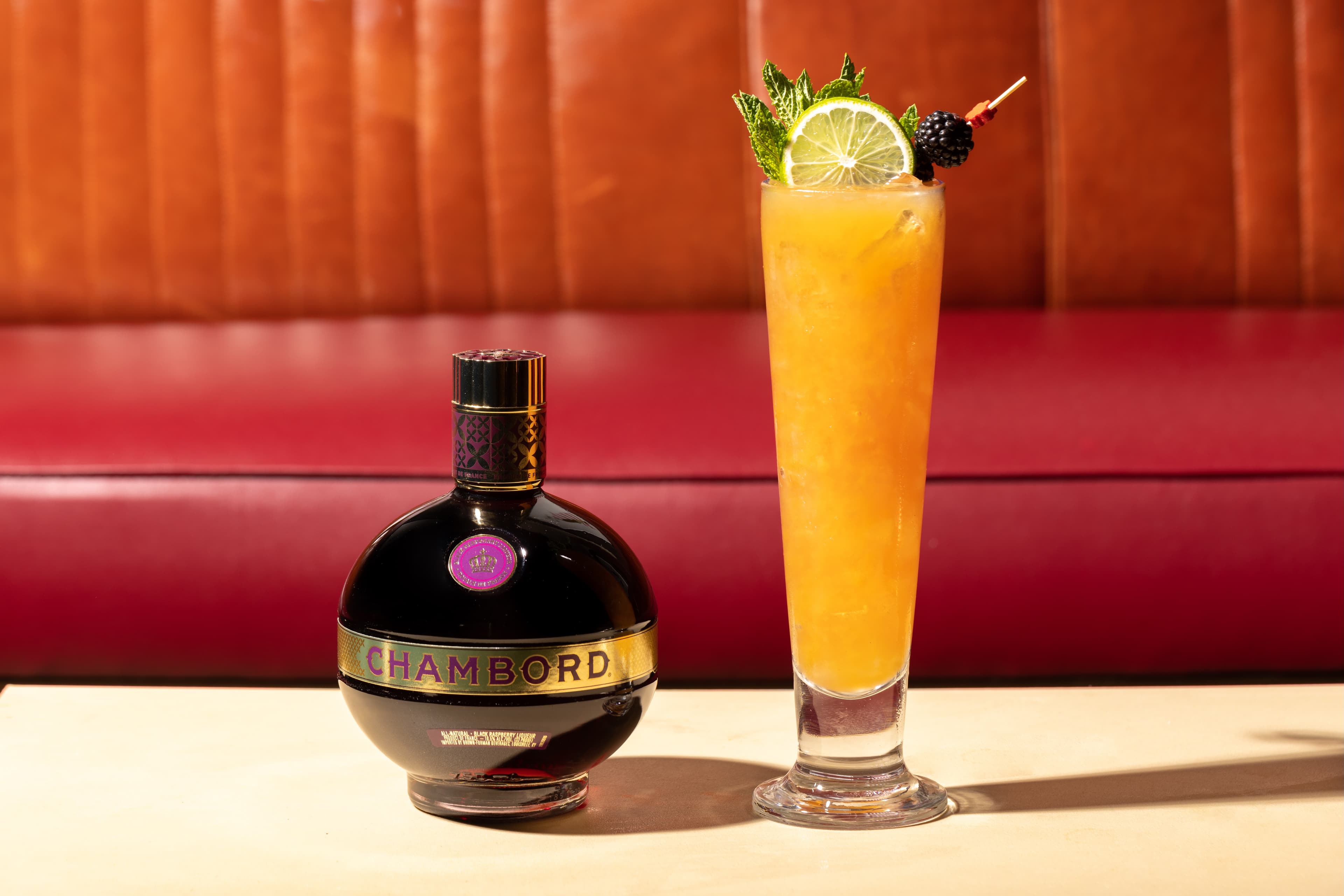 Chambord bottle beside a tall orange cocktail with mint and blackberry